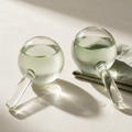 A pair of clear glass Lyquora Cryo Globes filled with pale sage liquid, misted with condensation on bone-white plaster